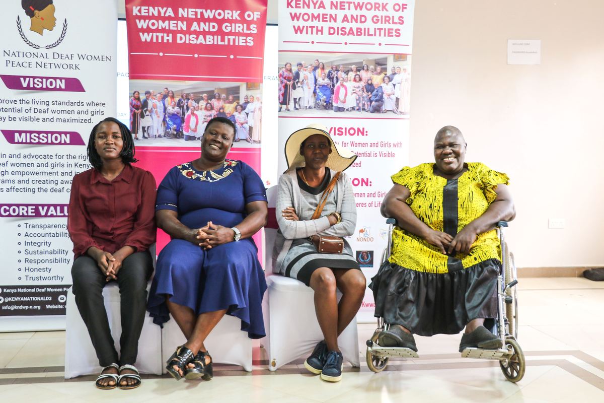 Four women, one using a wheelchair, seated in front of Kenya Network of Women and Girls with Disabilities banners